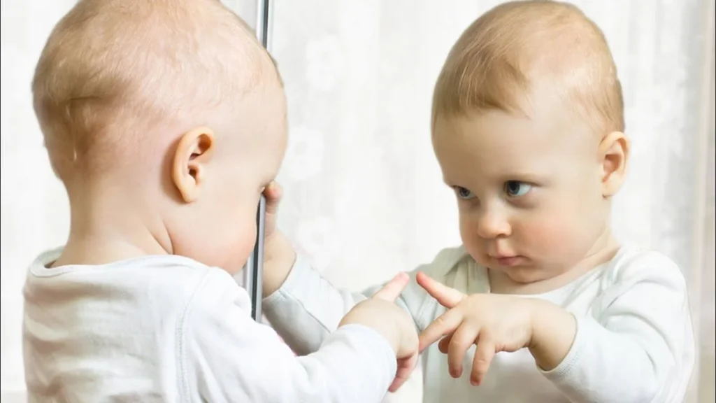 Baby pulling a funny face in front of a mirror and laughing at their reflection.
